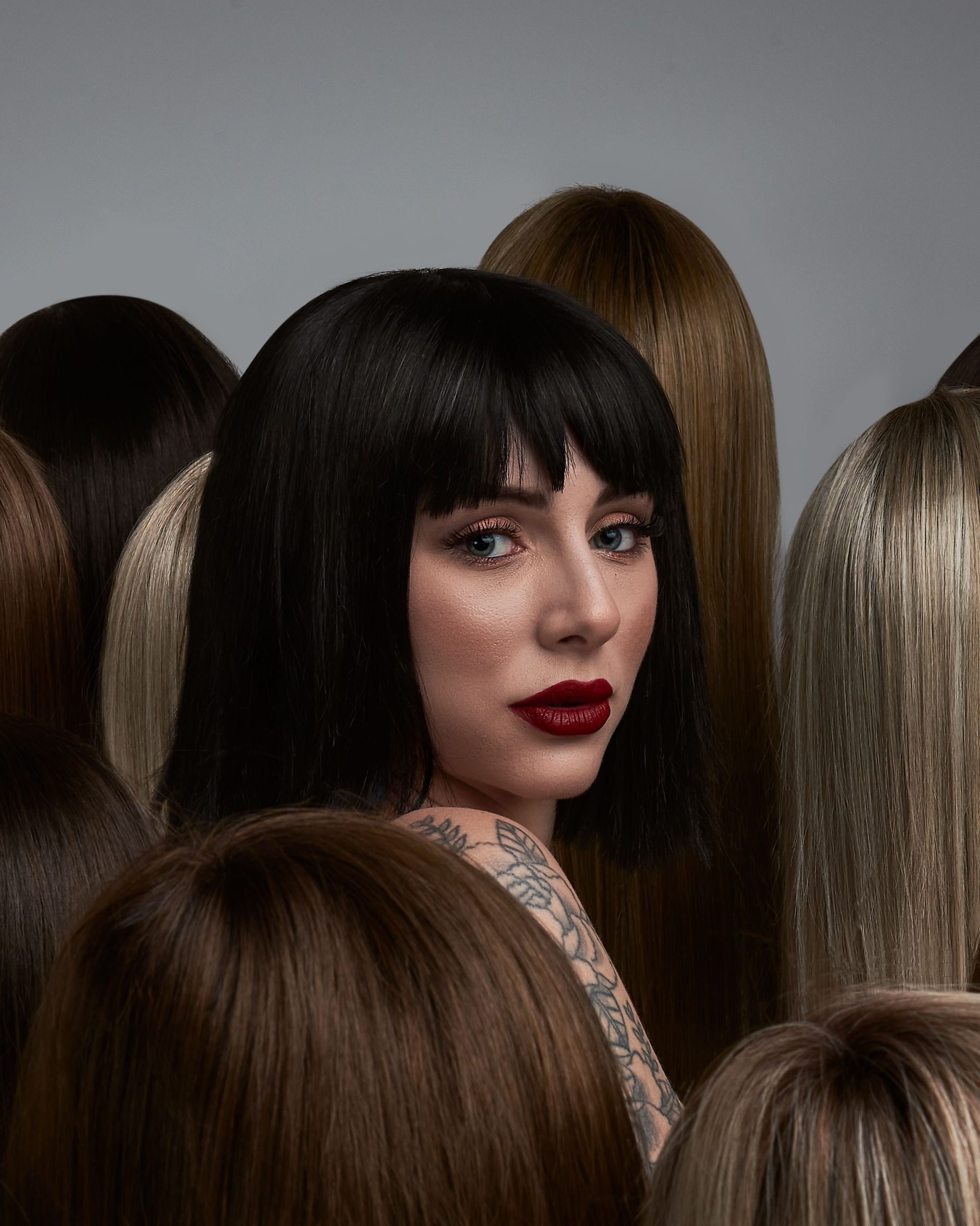 A striking portrait of a woman with dark hair amidst mannequin heads, showcasing diverse hairstyles.