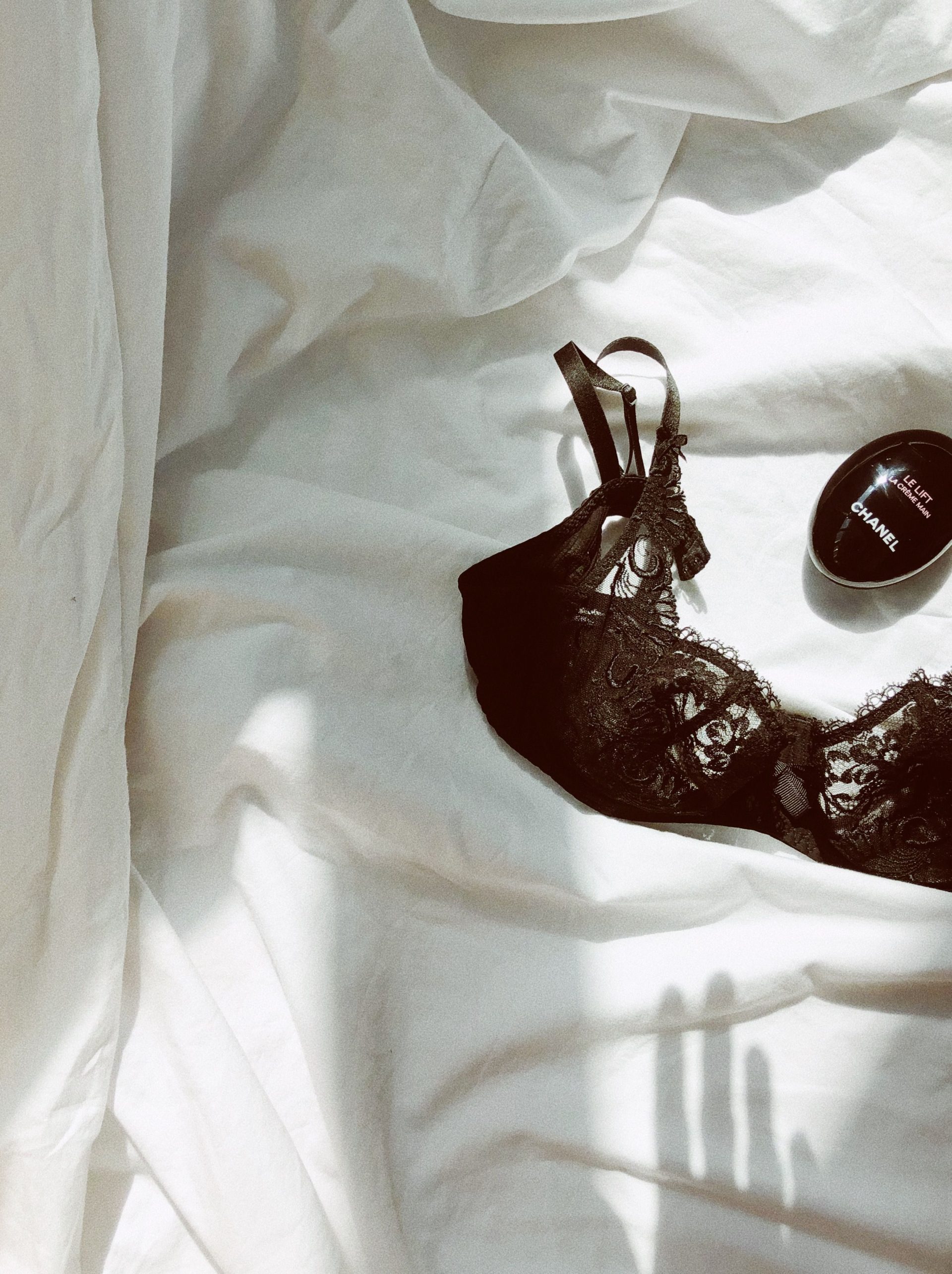 A stylish black lace bra and Chanel beauty product elegantly placed on soft white bedding under natural light.